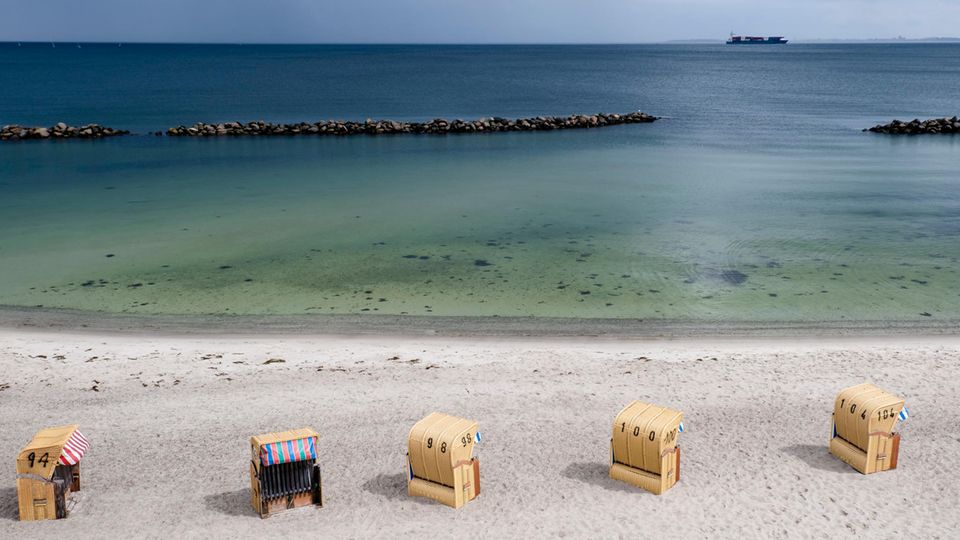 An einem Ostseestrand stehen fünf Strandkörbe in einer Reihe, während am Horizont ein Conatainerschiff vorbeifährt