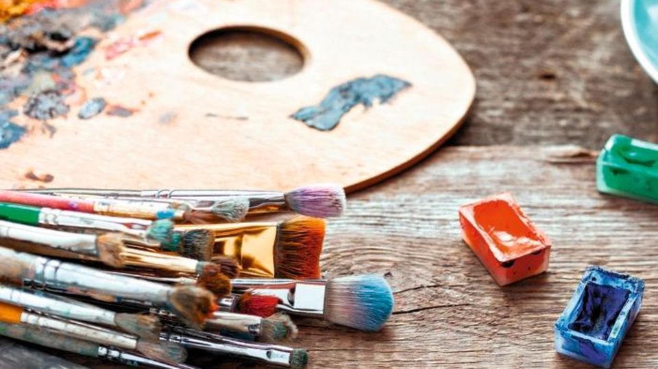 Paintbrushes, artist palette, pencils, coffee cup, watercolor and oil paints on desk in painter studio. Allerlei Malutensilien