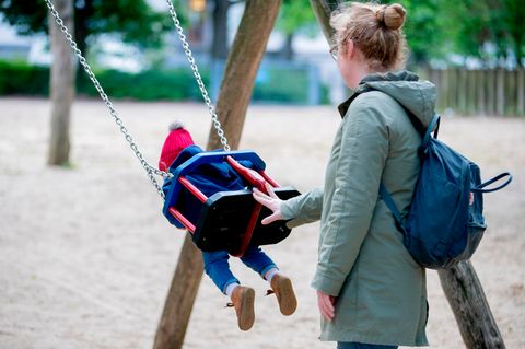 Eine Mutter in graugrünem Parka schubst auf einem Spielplatz ein Kind mit roter Mütze auf der Schaukel an