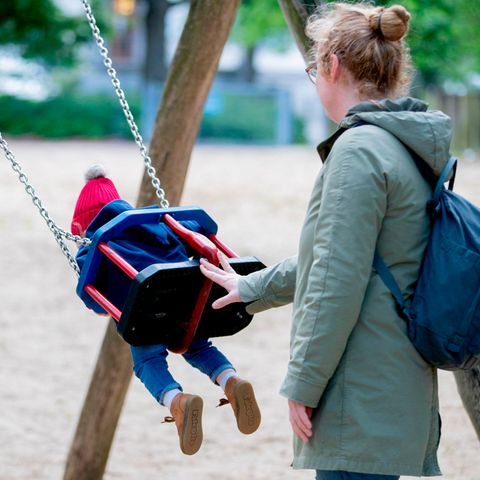 Eine Mutter in graugrünem Parka schubst auf einem Spielplatz ein Kind mit roter Mütze auf der Schaukel an