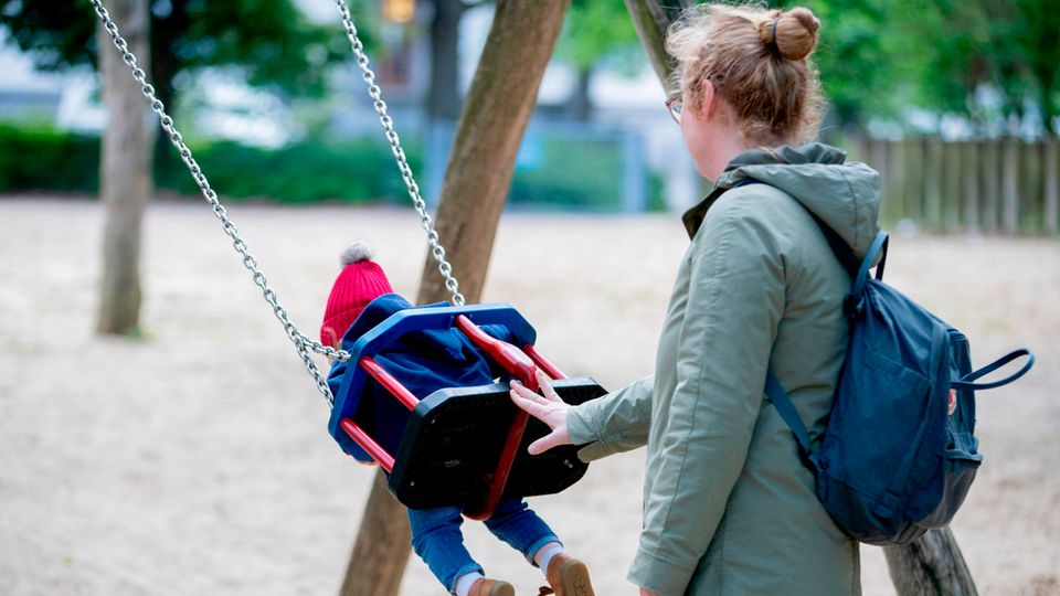 Eine Mutter in graugrünem Parka schubst auf einem Spielplatz ein Kind mit roter Mütze auf der Schaukel an