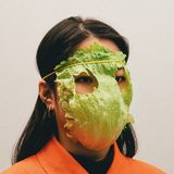 Eine Frau trägt ein Salatblatt mit Augenlöchern vor dem Gesicht