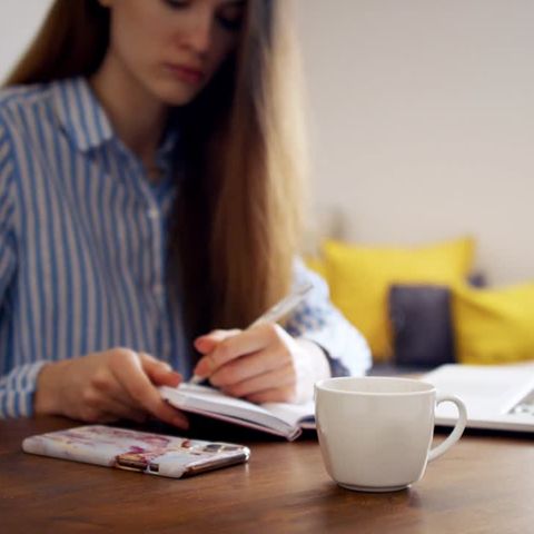 Eine langhaarige Frau sitzt an einem Schreibtisch mit Laptop und notiert sich etwas in einen Kalender