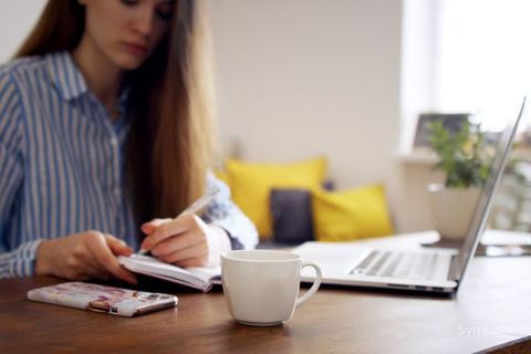 Eine langhaarige Frau sitzt an einem Schreibtisch mit Laptop und notiert sich etwas in einen Kalender