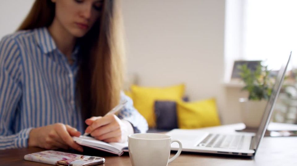 Eine langhaarige Frau sitzt an einem Schreibtisch mit Laptop und notiert sich etwas in einen Kalender