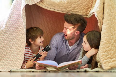 Vater und zwei Kinder mit Taschenlampe beim Buch vorlesen unter einer Decke