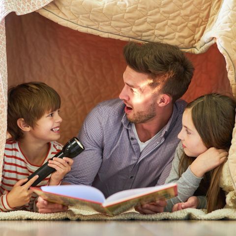 Vater und zwei Kinder mit Taschenlampe beim Buch vorlesen unter einer Decke