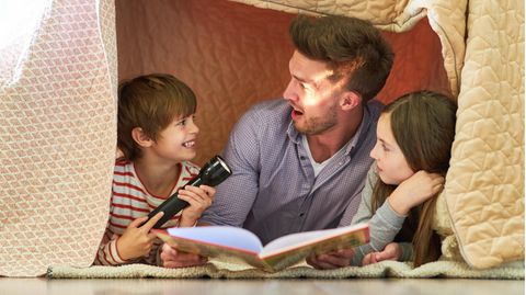 Vater und zwei Kinder mit Taschenlampe beim Buch vorlesen unter einer Decke
