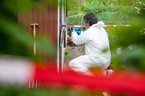 Ein Mann in weißem Overall kniet vor einer Gartenlaube und bohrt mit einer Bohrmaschine offenbar die Tür auf