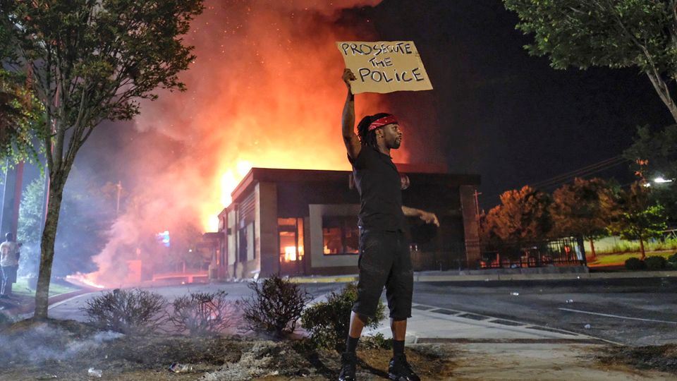 Schnellrestaurant in Flammen: Ein Mann hält ein Schild, während im Hintergrund das Wendy's-Restaurant in Atlanta brennt. Ein Mann hält ein Schild, während im Hintergrund das Wendy's-Restaurant in Atlanta brennt.