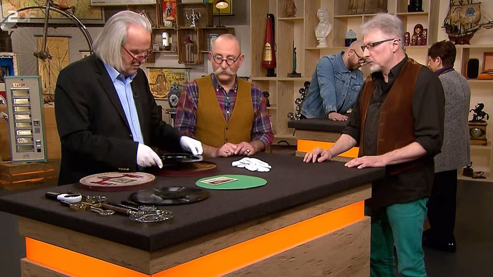 Bares für Rares "Bares für Rares"-Experte Albert Maier nimmt eine alte Schellackplatte in Augenschein. Moderator Horst Lichter und Verkäufer Horst Henry Schmitt hören seinen Ausführungen zu.