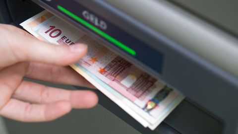 Eine linke Hand greift nach einem Euro-Scheinen, die aus dem Schacht eines Geldautomaten ragen