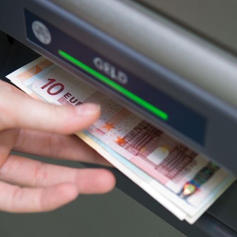 Eine linke Hand greift nach einem Euro-Scheinen, die aus dem Schacht eines Geldautomaten ragen