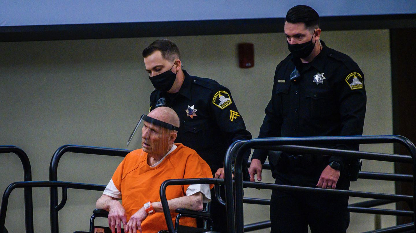 Ein Polizist fährt einen kahlköpfigen Mann in Rollstuhl und orangener US-Häftlingskleidung eine Rampe hinauf