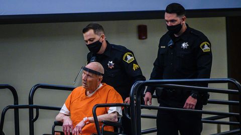 Ein Polizist fährt einen kahlköpfigen Mann in Rollstuhl und orangener US-Häftlingskleidung eine Rampe hinauf