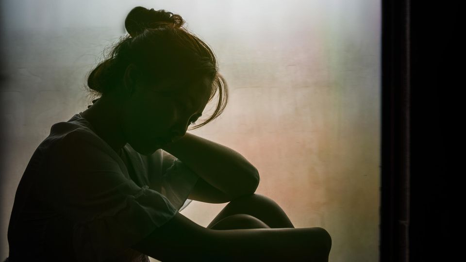 Einsamkeit in Lockdown Junge Frau sitzt im Schatten vor einem Fenster und umschlingt ihre Beine mit ihren Armen
