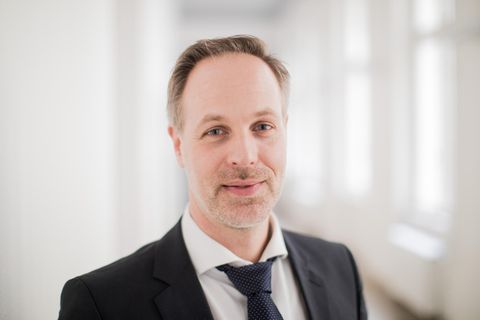 Ein Mann in schwarzem Jackett lächelt mit grau-blonden Haaren und Dreitagebart in die Kamera. Er hat blaue Augen