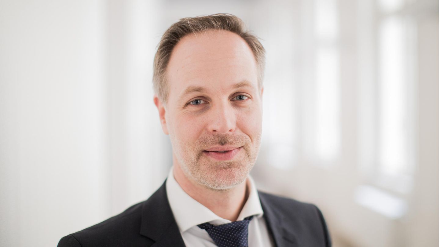 Ein Mann in schwarzem Jackett lächelt mit grau-blonden Haaren und Dreitagebart in die Kamera. Er hat blaue Augen