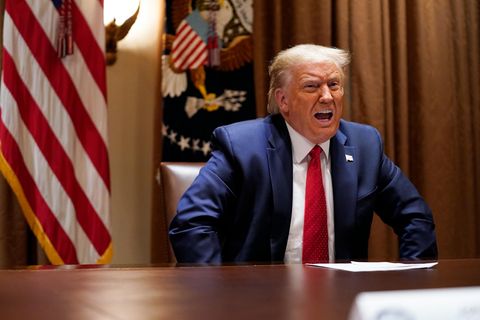 Us-Präsident Donald Trump sitzt in Anzug und Krawatte vor US-Flaggen an einem runden Tisch und spricht