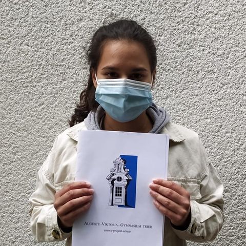 Eine junge Frau mit schwarzem Zopf steht mit Mundschutz vor einer verputzen Wand und hält sich ihr Abizeugnis vor die Brust