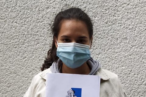Eine junge Frau mit schwarzem Zopf steht mit Mundschutz vor einer verputzen Wand und hält sich ihr Abizeugnis vor die Brust