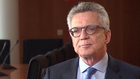 Thomasd de Maiziere sitzt in blauem Anzug und Krawatte neben einem Konferenztisch mit vielen Mikrofonen