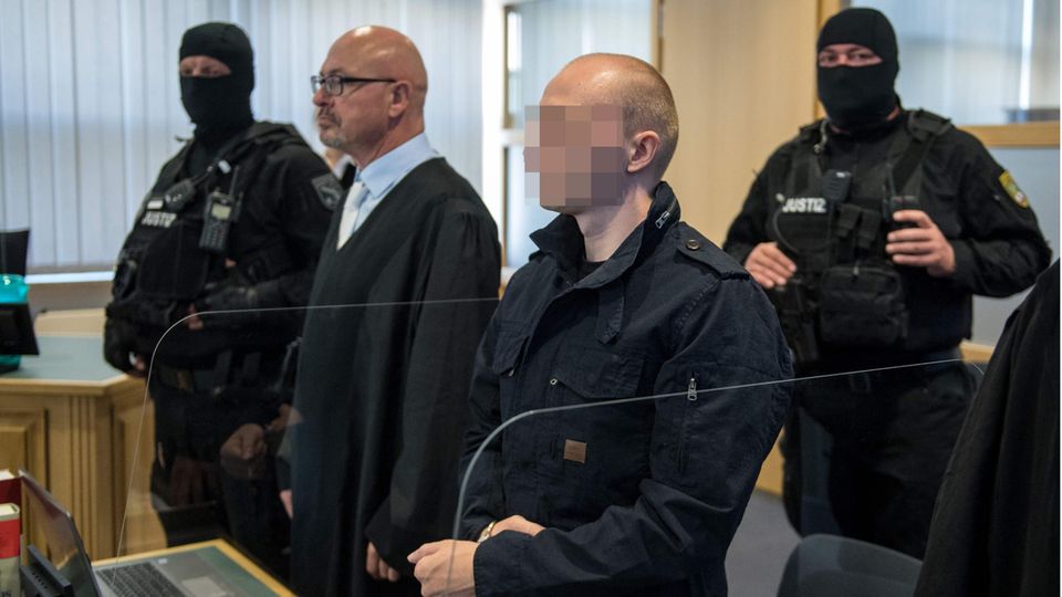 Der Angeklagte im Halle-Prozess im Gerichtssaal mit Anwalt und zwei Polizisten