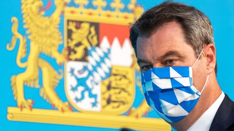 Bayerns Ministerpräsident Markus Söder mit weiß-blauer Gesichtsmaske vor bayerischem Wappen