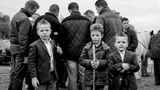 Drei Jungen mit Stöcken stehen vor einer Gruppe Männer