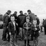 Drei Jungen mit Stöcken stehen vor einer Gruppe Männer