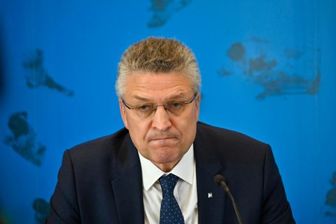 Ein Mann mit grau melierten Locken und randloser Brille sitzt in Anzug und Krawatte vor einer blauen Wand mit Weltkarte