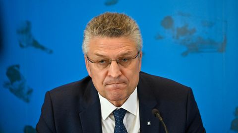 Ein Mann mit grau melierten Locken und randloser Brille sitzt in Anzug und Krawatte vor einer blauen Wand mit Weltkarte