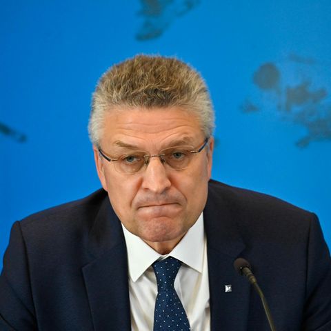 Ein Mann mit grau melierten Locken und randloser Brille sitzt in Anzug und Krawatte vor einer blauen Wand mit Weltkarte
