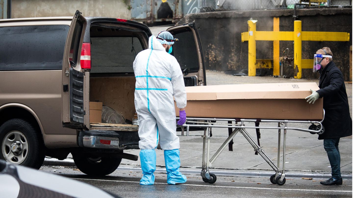 Zwei Menschen in Schutzkleidung fahren einen Sarg auf einem Gestell an die offenen Hecktüren eines Vans heran