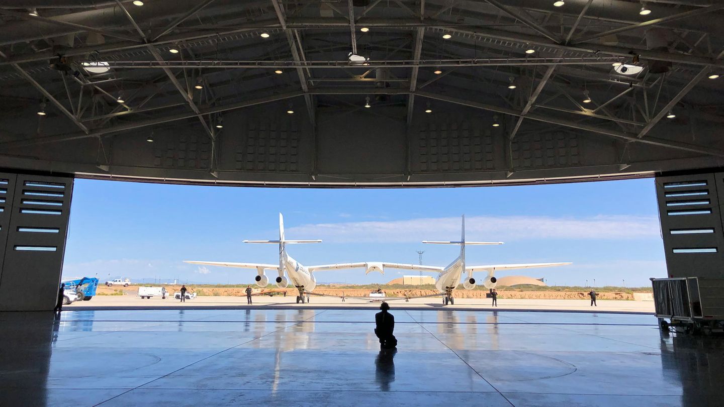 Vor dem Hangar steht das doppelrümpfige Trägerflugzeug. In der Mitte wird der Raumgleiter mit dem Namen "VSS Unity" befestigt. VSS steht für Virgin Space Ship.