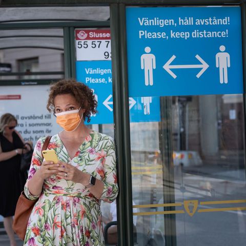 Eine Frau mit Mund-Nase-Schutz wartet an einer Bushaltestelle in Stockholm. Ein Schild weist auf Abstandsregeln hin.