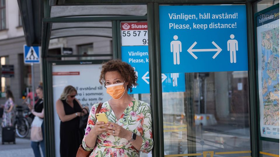 Eine Frau mit Mund-Nase-Schutz wartet an einer Bushaltestelle in Stockholm. Ein Schild weist auf Abstandsregeln hin.