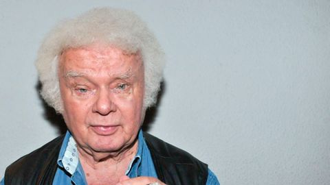 Ein Mann mit weißem Minipli hält eine Zigarre in der linken Hand und schaut gerade in die Kamera