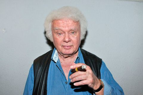 Ein Mann mit weißem Minipli hält eine Zigarre in der linken Hand und schaut gerade in die Kamera