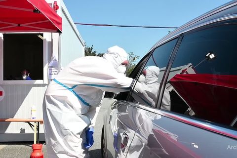 Ein Mann in weißem Schutz-Overall beugt sich an das offene Fahrerfenster eines Autos, um einen Coronatest zu machen