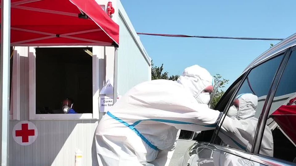 Coronatest Bayern Auto Ein Mann in weißem Schutz-Overall beugt sich an das offene Fahrerfenster eines Autos, um einen Coronatest zu machen