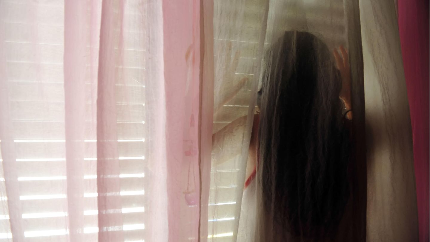 Junges Mädchen steht hinter einer Gardine vor einem Fenster mit einer Jalousie