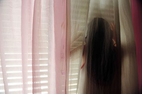Junges Mädchen steht hinter einer Gardine vor einem Fenster mit einer Jalousie