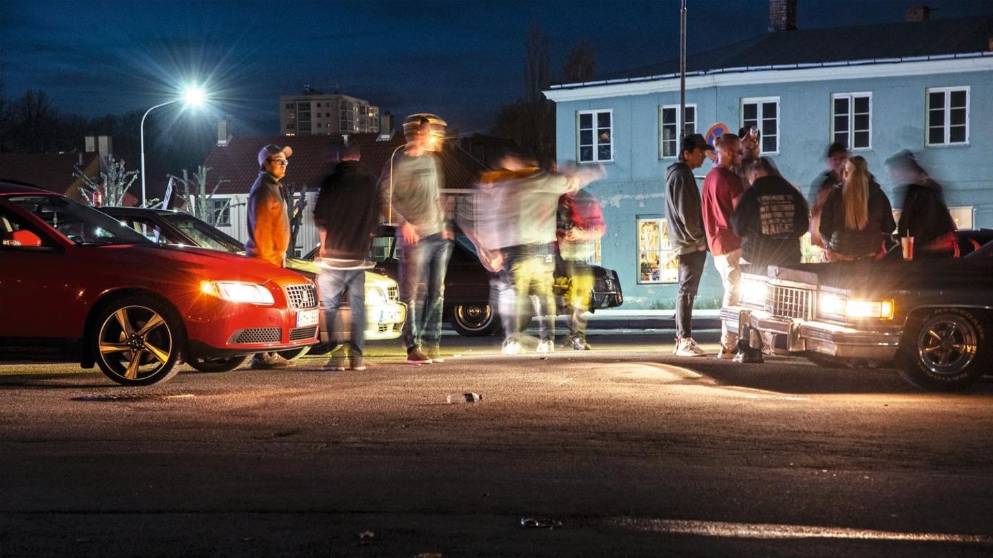 Stadtzentrum von Halden, Herumstehen, dazukommen, ein paar Runden fahren, wieder dastehen, mit Freunden abhängen. Jung sein. Die Straße, der Parkplatz, die Autos sind sozialer Treffpunkt, in dem Gemeinschaft, Freundschaft und Zugehörigkeit entsteht und ... Zwei Autos leuchten eine Gruppe von Menschen an
