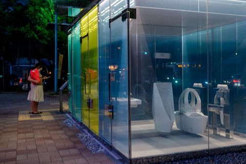 Eine Frau in Rock und rotem Oberteil steht vor einem Toilettenhäuschen, durch dessen Glaswand man eine Toilettenschüssel sieht