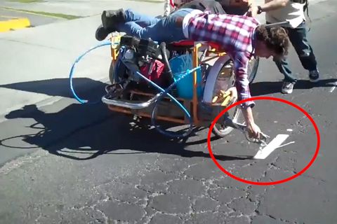 Ein Mann im Karohemd liegt auf einem Lastenfahrrad und malt weiße Markierungen auf die Fahrbahn