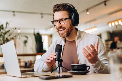 Glücklich lächelnder bärtiger Mann mittleren Alters, der Podcast im Café mit Laptop und Mikrofon aufnimmt