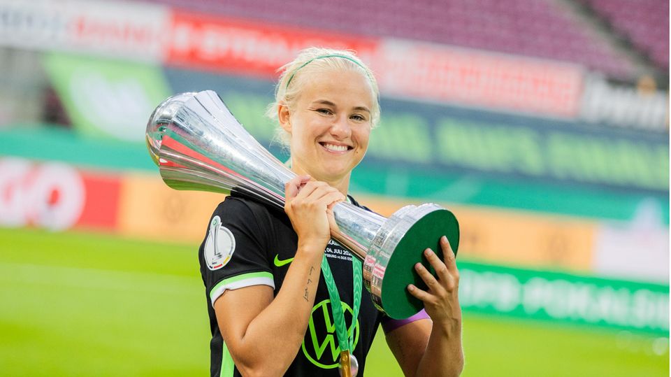 Eine blonde Frau mit Haarreif trägt das dunkelgrüne Trikot des VfL Wolfsburg und stemmt mit beiden Händen einen silbernen Pokal