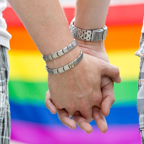 Die Hände zweier Männer, ineinander verschränkt, vor Regenbogenfahne