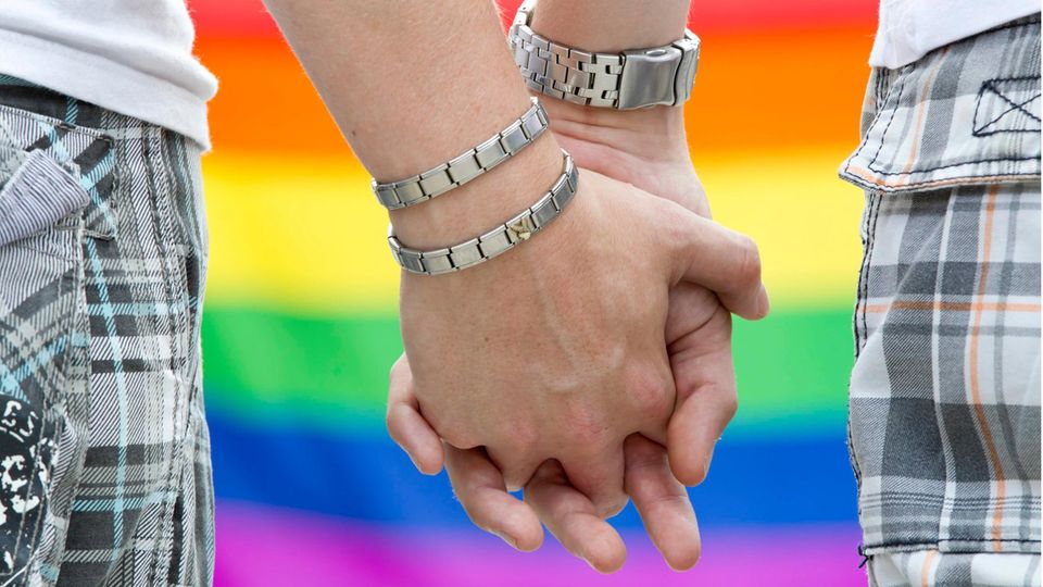 Die Hände zweier Männer, ineinander verschränkt, vor Regenbogenfahne